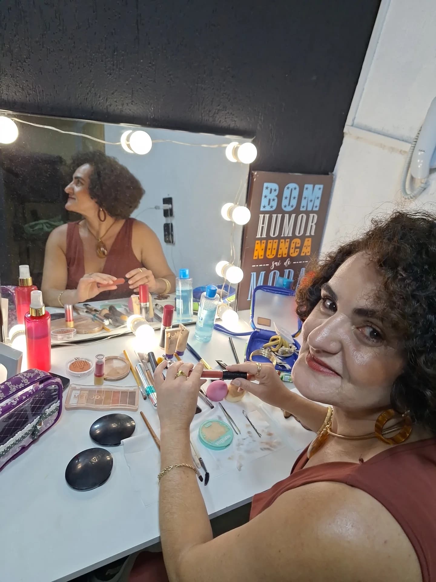 Nellva segurando um batom diante de um espelho iluminado e uma bancada de maquiagem