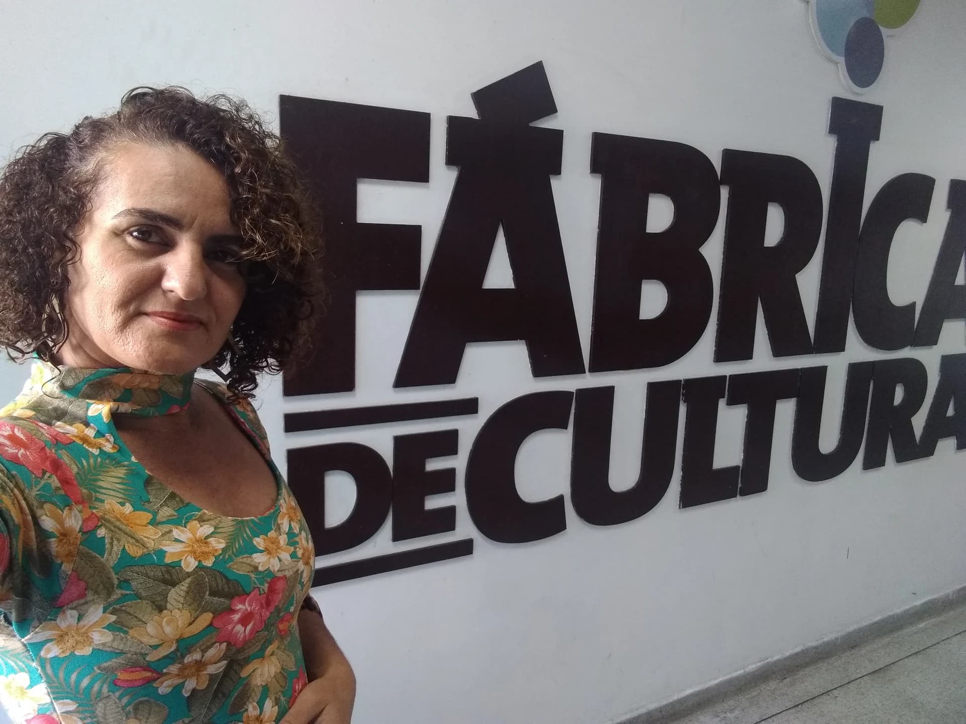 Nellva de blusa floral diante da placa Fábrica de Cultura