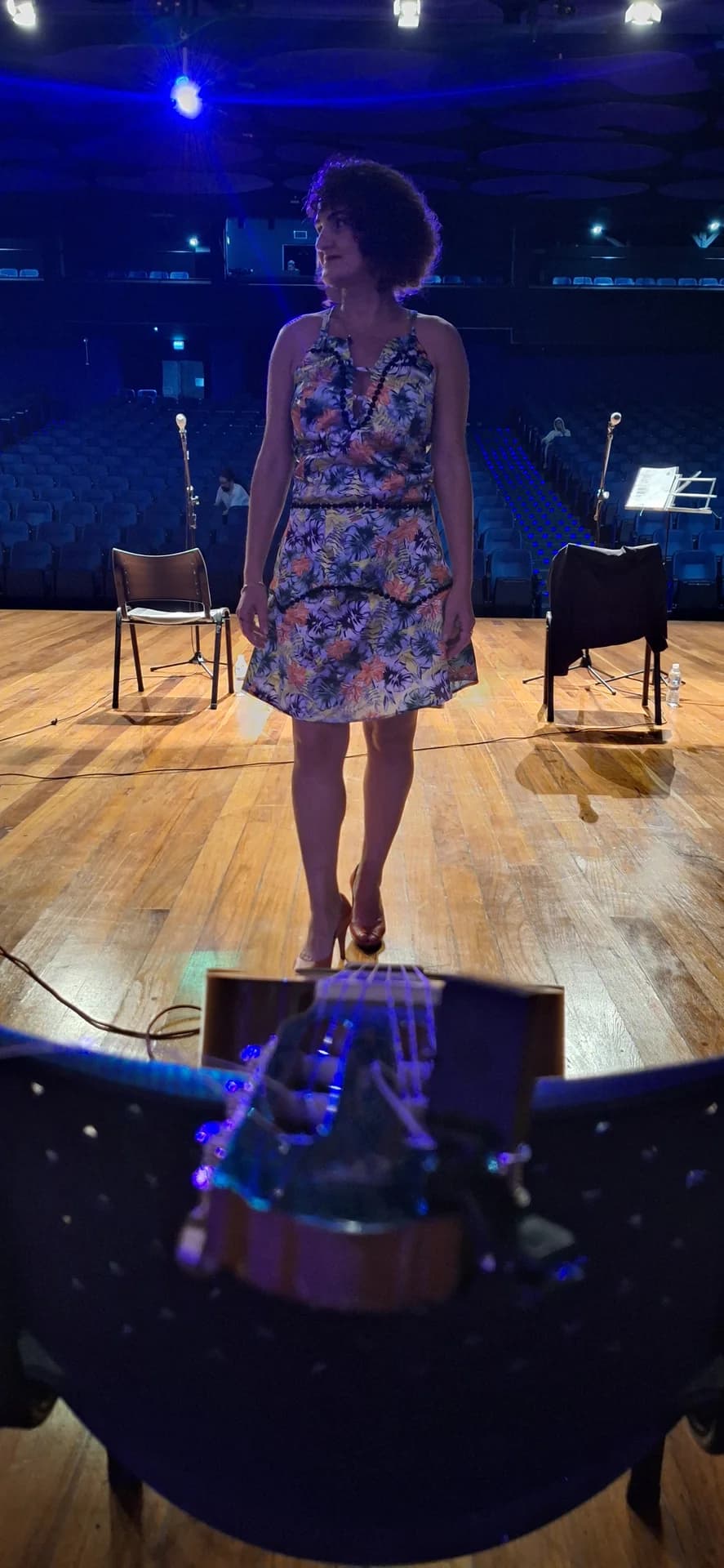 Nellva de vestido floral sobre um palco, com cadeiras da plateia ao fundo
