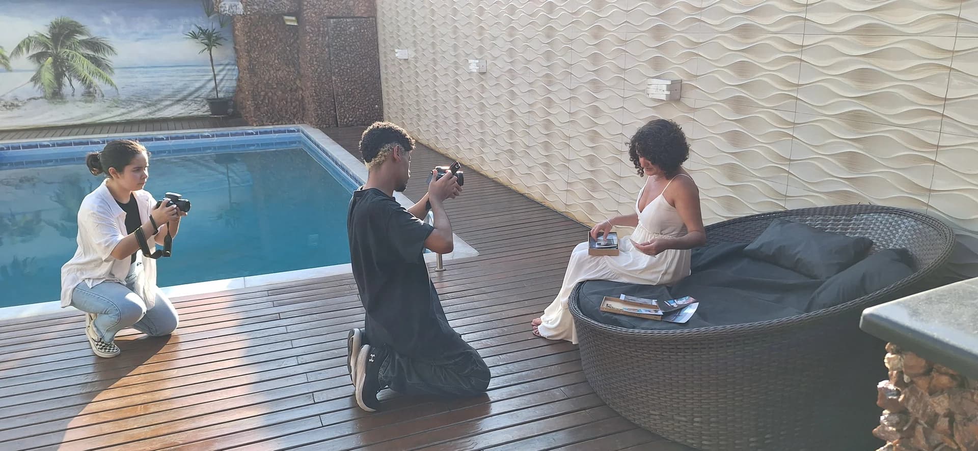 Nellva sentada com fotografias nas mãos, diante de duas pessoas com câmeras junto a uma piscina