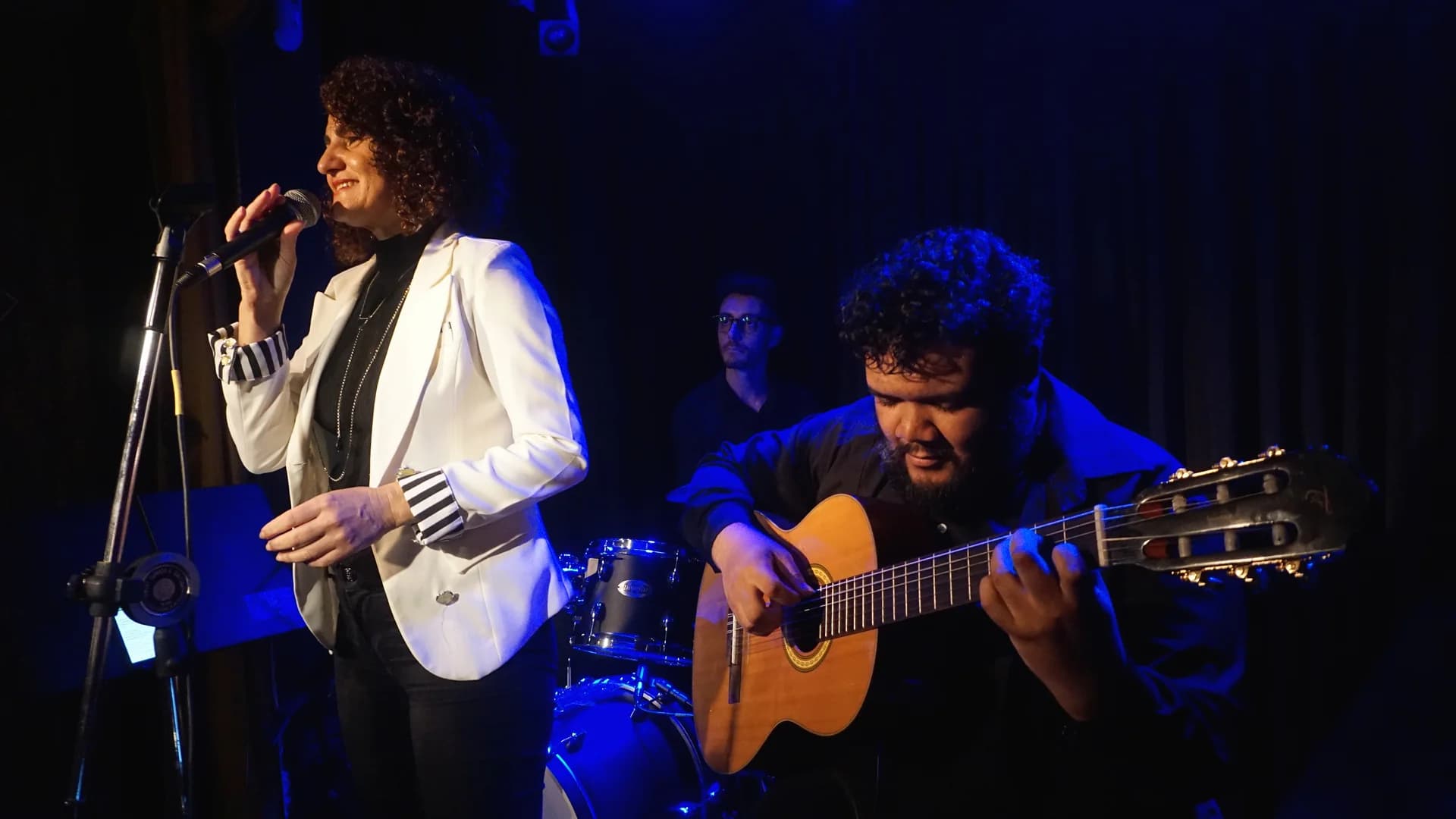 Nellva de blazer branco cantando ao lado de um violonista sob luz azul