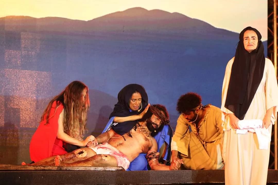 Nellva de véu escuro e roupa clara à direita de uma cena teatral com outros intérpretes