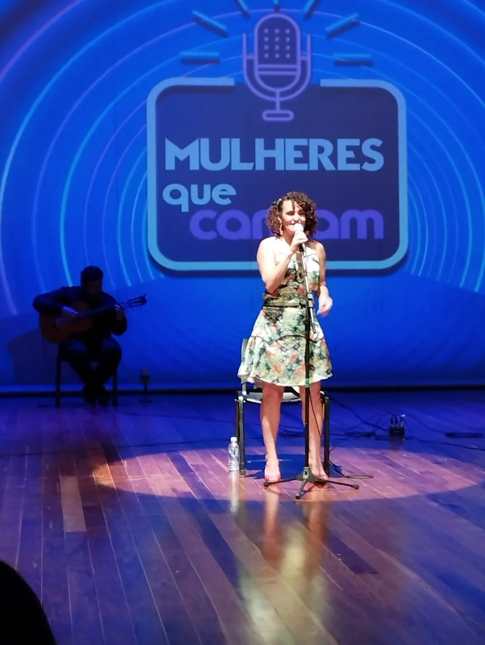 Nellva de vestido floral cantando diante de uma projeção azul