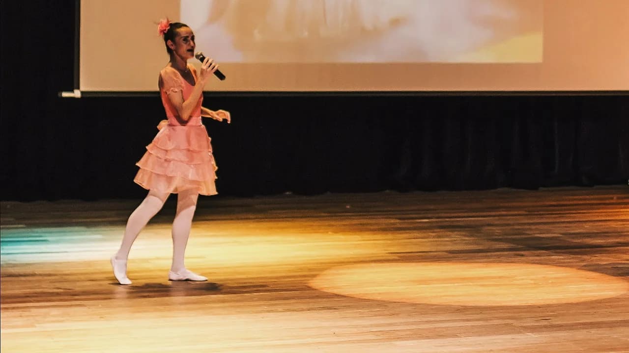Nellva cantando com vestido rosa e sapatilhas em um palco de madeira