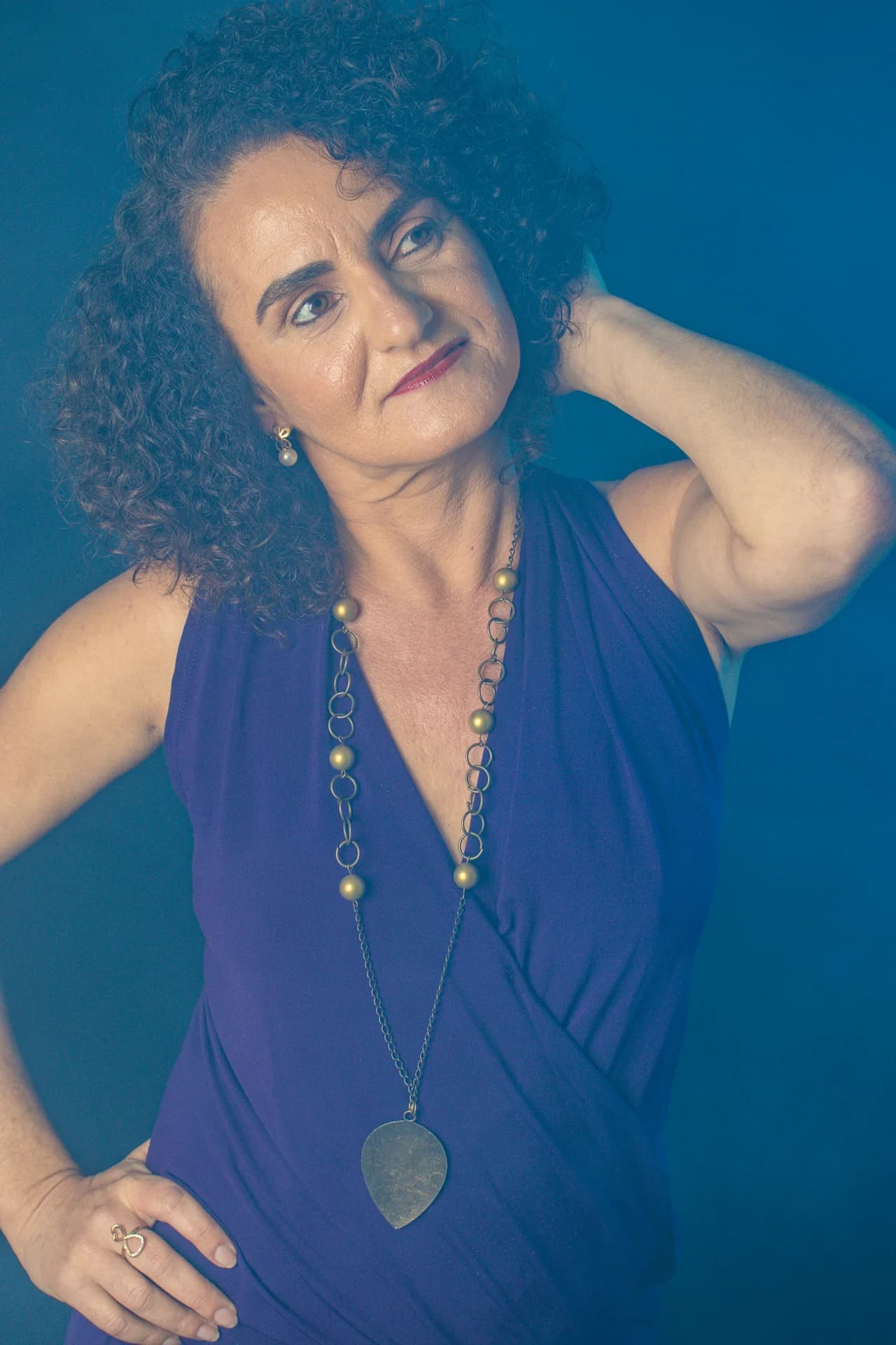 Nellva de blusa azul com uma mão no cabelo e colar longo