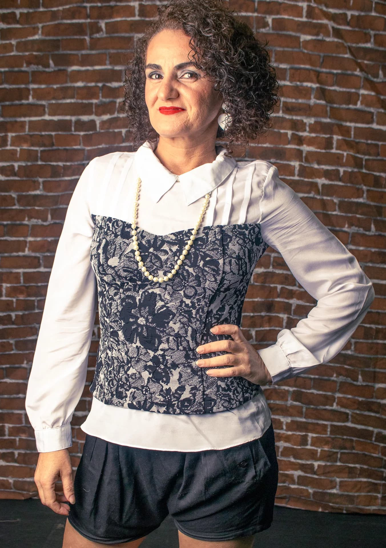 Nellva de camisa branca e renda escura diante de uma parede de tijolos